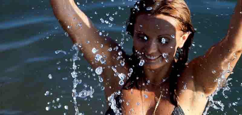 Eine Frau im Bikini steht fröhlich in einem Gewässer, während sie das Wasser aufwirbelt und spritzt, umgeben von funkelnden Wassertropfen und Sonnenlicht.